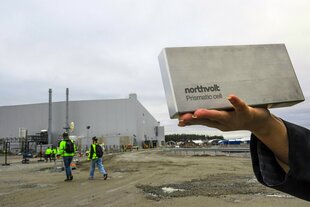 Ein Hand mit Northvolt-Werbeblock und die Fabrik in Skellefteå im Hintergrund