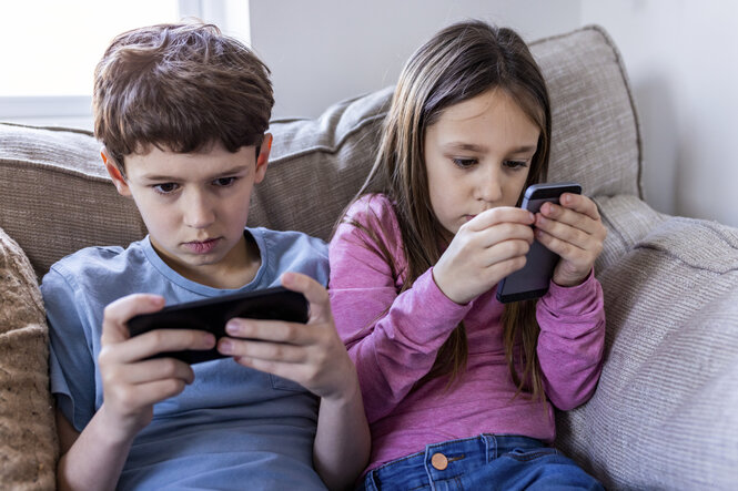 2 Kinder auf Sofa, auf Smartphones blickend