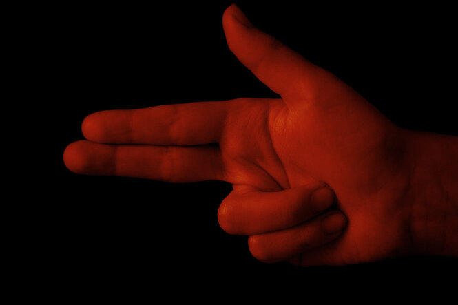 Eine Hand deutet mit zwei Fingern eine Schusswaffe an, sie ist in dunkelrotes Licht getaucht
