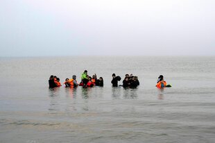 Frauen, Kinder und Männer stehen bis zur Hüfte im Wasser, manche von ihnen tragen Rettungswesten