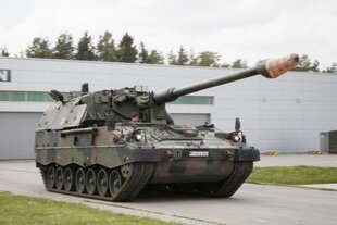 Zu sehen ist ein Raupenpanzer der Bundeswehr, vor einem grauen Gebäude mit Garagentoren, der Kopf eines Soldaten schaut aus dem Gefährt heraus.