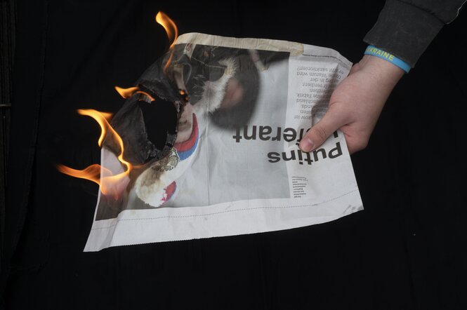 Eine brennende Zeitung wird vor einem schwarzem Hintergrund mit einer Hand gehalten.