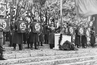 NS-Propaganda­minister Joseph Goebbels steht gestikulierend an einem Rednerpult. Um ihn herum stehen uniformierte Fahnenträger mit Hakenkreuzfahnen.