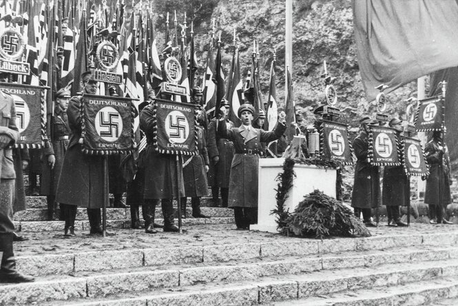 NS-Propaganda­minister Joseph Goebbels steht gestikulierend an einem Rednerpult. Um ihn herum stehen uniformierte Fahnenträger mit Hakenkreuzfahnen.