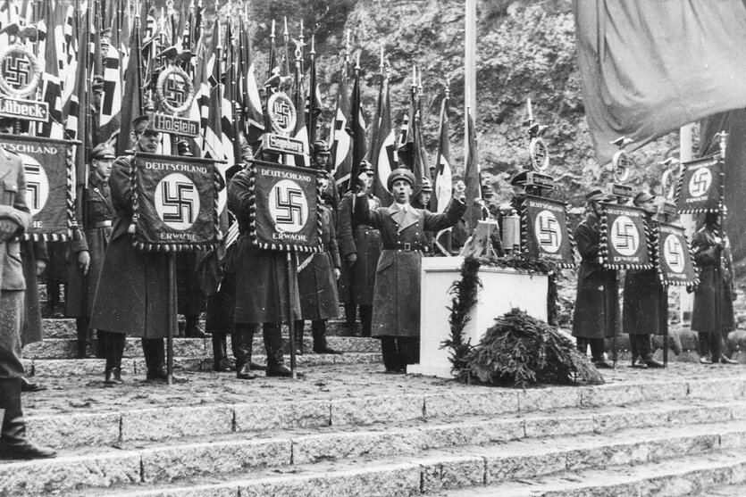 NS-Propaganda­minister Joseph Goebbels steht gestikulierend an einem Rednerpult. Um ihn herum stehen uniformierte Fahnenträger mit Hakenkreuzfahnen.