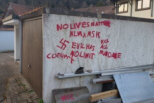 Auf der Wand einer Garage sind mit roter Farbe mehrere Graffitis zu sehen, darunter ein Hakenkreuz und Schriftzüge mit gewaltverherrlichenden und menschenfeindlichen Aussagen. Einige der Begriffe sind verschlüsselt oder kryptisch. Vor der Garage liegen Metallplatten und Bauschutt.
