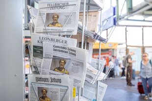 In einem Bahnhof steht ein Zeitungsständer mit verschiedenen regionalen Tageszeitungen, deren Titelblätter größtenteils das gleiche Motiv zeigen – eine historische Skulptur mit der Überschrift „Deutscher Wahrheitssucher“.