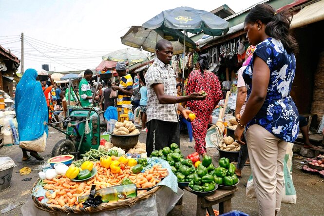 Markt in LAgos, ein Händler zeigt einer Kundin seine Paprika, im Hintergrund sind weitere Marktstände und einkaufende Menschen in bunter Kleidung zu sehen