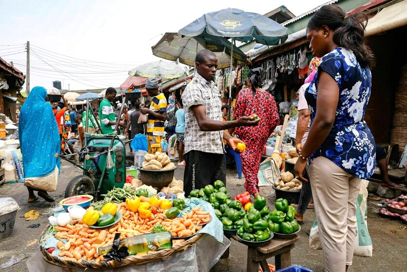 Markt in LAgos, ein Händler zeigt einer Kundin seine Paprika, im Hintergrund sind weitere Marktstände und einkaufende Menschen in bunter Kleidung zu sehen