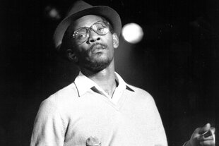 Linton Kwesi Johnson mit Hut und Bbrille, in der Hand ein Mikrofon, auf einer Konzertbühne