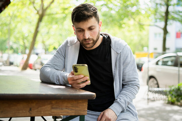 Wael Aljasim, Geflüchteter aus Syrien sitzt am Tisch eines Straßencafes und schaut auf sein Smartphone