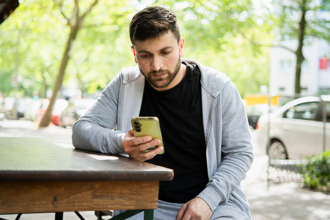 Wael Aljasim, Geflüchteter aus Syrien sitzt am Tisch eines Straßencafes und schaut auf sein Smartphone