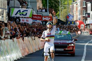 Ein Fahrer auf dem Rad reißt vor Publikum jubelt einen Arm hoch und zeigt gen Himmel