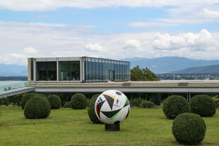 Ein Glasbau vor Bergpanorama, im Vordergrund ein Park mit einem riesigen Fußball