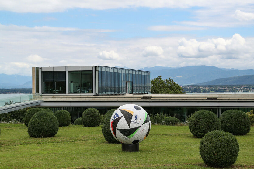 Ein Glasbau vor Bergpanorama, im Vordergrund ein Park mit einem riesigen Fußball
