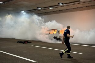 Ein Mann läuft an einem brennenden Auto vorbei, weißer Rauch ist sichtbar