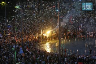 Menschen schwenken ihre Handys mit blinkenden Lichtern bei einer großen Kundgebung. Ein Bengalo erleuchtet eine serbische Flagge