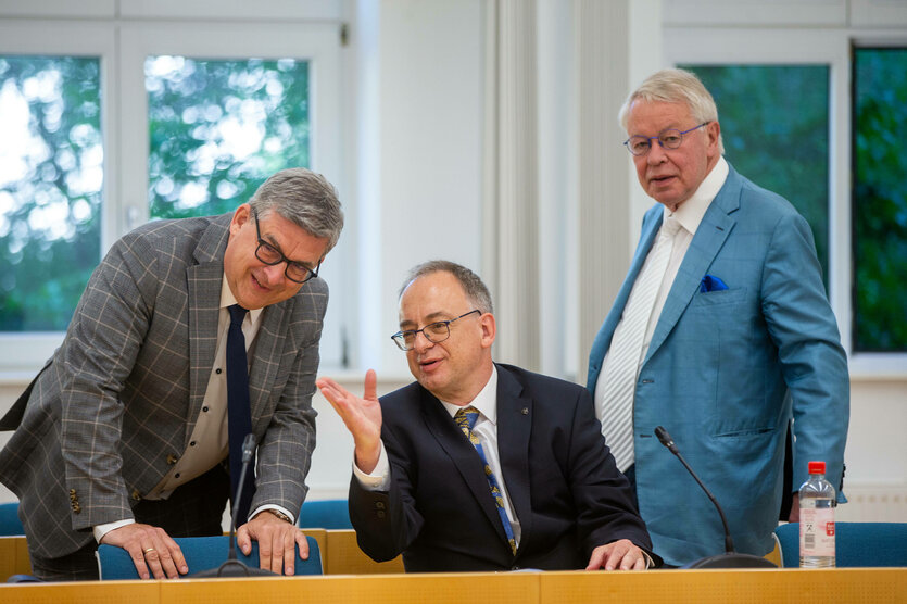 Drei Männer im Gerichtssaal, einer sitzt, die beiden anderen Männer stehen neben ihm und hören ihm zu