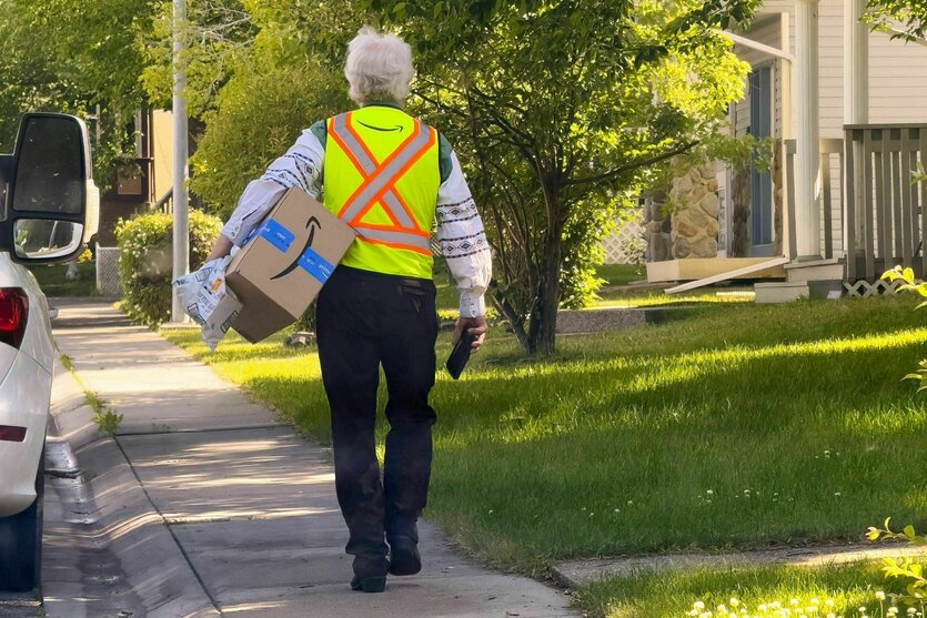 Ein älterer Mann mit Warnweste bekleidet trägt ein Paket mit dem Amazonlogo und geht eine Wohnstraße entlang