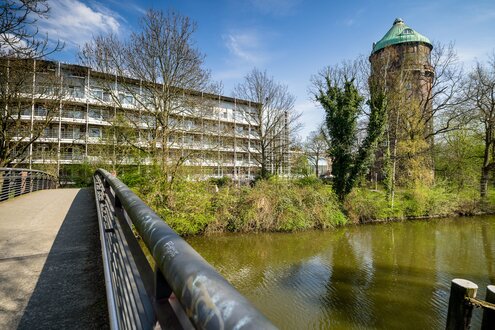 ein Bettenhaus und ein Wasserturm an einem Gewässer, über das eine Brücke führt