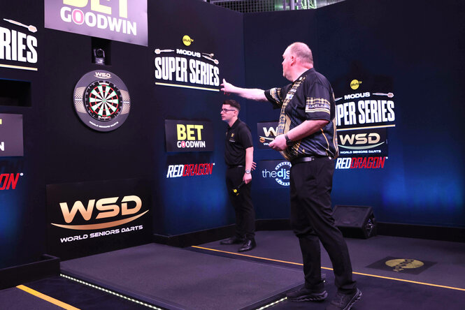 Darts-Turnier im englischen Portsmouth, Juni 2025 - Ein Mann wirft einen Pfeil Richtung Scheibe, das wird von einem weiteren Mann, der neben der Scheibe steht, beobachtet