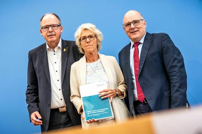 Christiane Schönefeld hält einen Bericht in die Kamera, die beiden Männer im Anzug stehen rechts und links neben ihr vor einer blauen Wand