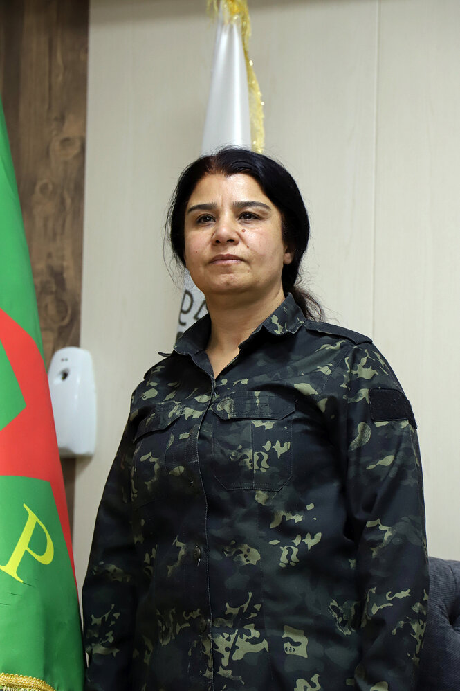 Nesrin Abdullah steht in militärischer Uniform neben einer Fahne des YPG