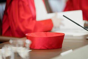 Die rote Kopfbedeckung eines Mitglieds des Bundesverfassungsgerichts liegt auf dem Tisch, im Hintergrund unscharf eine angeschnittene RichterIn