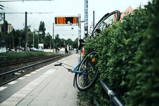 Ein schräg am Zaun einer Straßenbahnhaltestelle hängendens Fahrrad mit einem dichten Busch im Vordergrund. Dahinter befinden sich Straßenbahngleise, ein Bahnsteig und eine Anzeigetafel, auf der Abfahrtszeiten zu sehen sind. Im Hintergrund unspezifische S