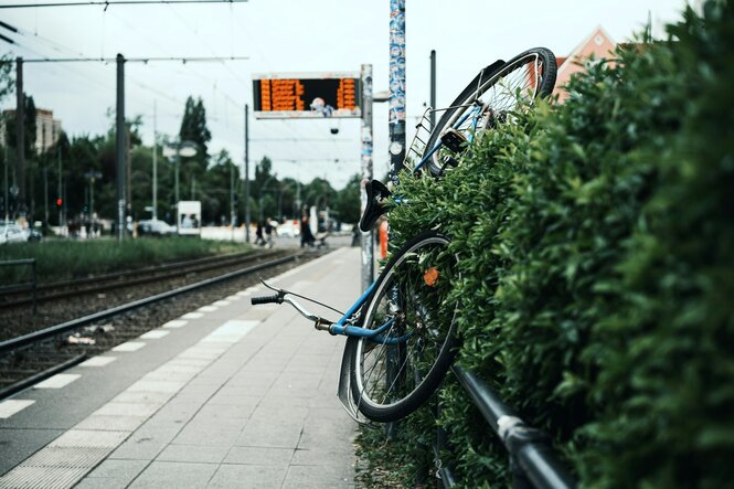 Ein schräg am Zaun einer Straßenbahnhaltestelle hängendens Fahrrad mit einem dichten Busch im Vordergrund. Dahinter befinden sich Straßenbahngleise, ein Bahnsteig und eine Anzeigetafel, auf der Abfahrtszeiten zu sehen sind. Im Hintergrund unspezifische Stadtlandschaft.