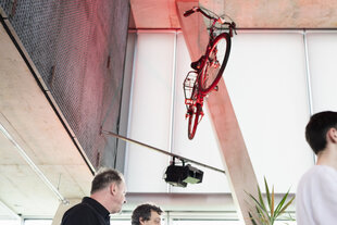 Das Fahrrad von Christian Ströbele hängt von der Decke im Neubau der taz. Das Fahrrad, beleuchtet von rotem Licht, ist in einer modernen, hohen Halle platziert, dem taz Panorama. Im Vordergrund betrachten zwei Männer das Andenken, während ein weiterer mit dem Rücken zum Betrachter steht. Eine Palme am rechten Bildrand fügt einen grünen Akzent hinzu.