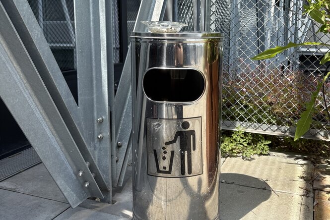 Chromener, hoher Mülleimer mit einem Piktogramm eines Menschen, der Müll entsorgt. Auf dem Mülleimer befindet sich ein gläserner Aschenbecher.