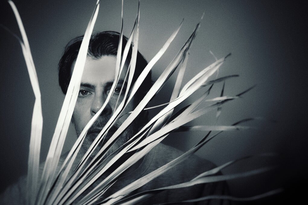 Schwarz-Weiß-Foto: Porträtbild des Musikers Andrew Pekler, er steht in einem Lichkegel, sein Gesicht schaut hinter Palmenblättern hervor, die ins Bild ragen.
