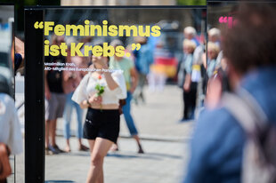 Der Satz „Feminismus ist Krebs“ steht auf einem Spiegel vor dem Brandenburger Tor, während Passantinnen und Passanten sich während des Lesens darin spiegeln. Der ist gekennzeichnet als Zitat des AfD-Politikers Maximilian Krah.