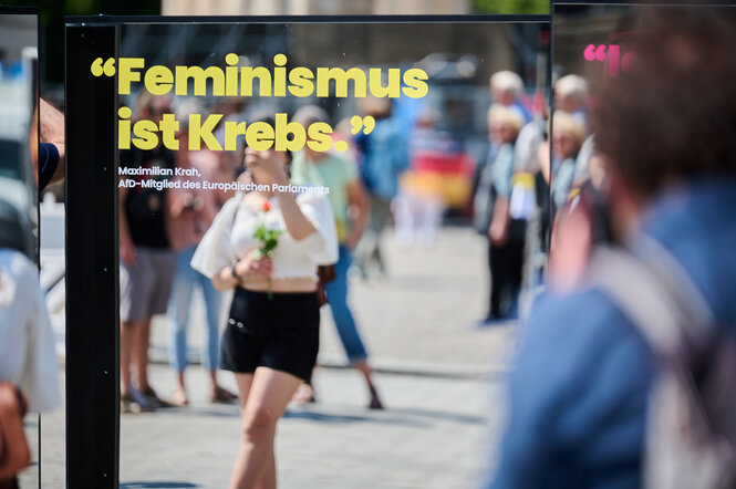 Der Satz „Feminismus ist Krebs“ steht auf einem Spiegel vor dem Brandenburger Tor, während Passantinnen und Passanten sich während des Lesens darin spiegeln. Der ist gekennzeichnet als Zitat des AfD-Politikers Maximilian Krah.