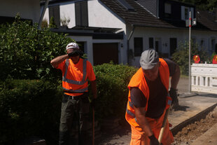 Zwei Straßenarbeiter in leuchtroter Arbeitskleider stehen auf einer Baustelle am Rand einer Straße, einer schirmt sein Gesicht gegen das Sonnenlicht ab