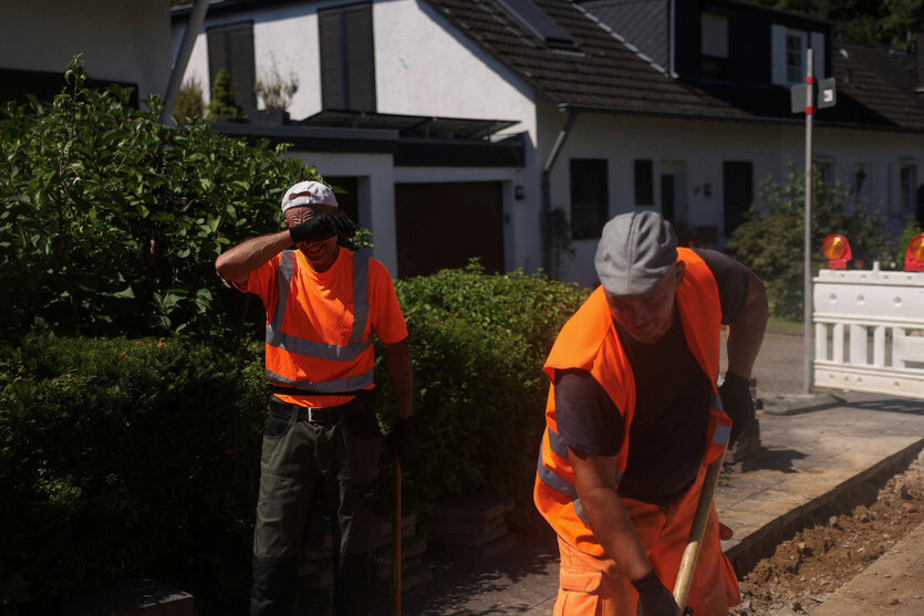 Zwei Straßenarbeiter in leuchtroter Arbeitskleider stehen auf einer Baustelle am Rand einer Straße, einer schirmt sein Gesicht gegen das Sonnenlicht ab