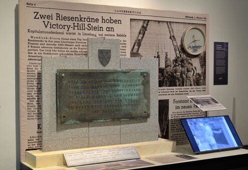 Eine Gedenktafel mit der Inschrift zur Kapitulation der deutschen Streitkräfte am 4. Mai 1945 steht vor einer alten Zeitung mit der Überschrift "Zwei Riesenkräne hoben Victory-Hill-Stein an". Im Hintergrund sind ein Zähler mit dem Datum 30. September 1958 und Fotos einer Baustelle zu sehen. Ein Bildschirm und weitere Ausstellungsstücke sind darunter angeordnet.