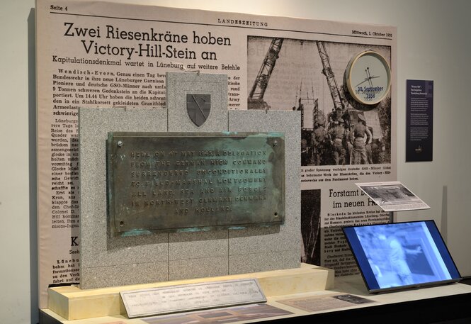 Eine Gedenktafel mit der Inschrift zur Kapitulation der deutschen Streitkräfte am 4. Mai 1945 steht vor einer alten Zeitung mit der Überschrift "Zwei Riesenkräne hoben Victory-Hill-Stein an". Im Hintergrund sind ein Zähler mit dem Datum 30. September 1958 und Fotos einer Baustelle zu sehen. Ein Bildschirm und weitere Ausstellungsstücke sind darunter angeordnet.