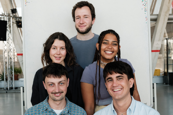 Das Bild vor einer großen weißen Pinnwand zeigt fünf junge Erwachsene, die pyramidenförmig posieren: zwei unten, zwei in der Mitte, eine Person oben. Alle blicken lächelnd oder schmunzelnd in die Kamera. Die Kleidung ist lässig, das Licht natürlich.