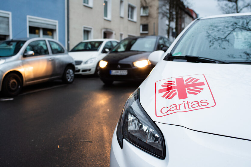 Ein weißes Auto mit Caritas-Logo ist am Rand einer Straße geparkt, in der Reihenhäuser, andere parkende Autos stehen und durch die ein schwarzes Auto mit Scheinwerfern fährt.