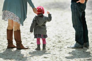 Ein kleines Mädchen steht mit seinen Eltern auf Sand - von den Eltern sind nur die Beine zu erkennen