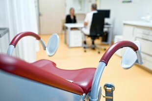 Gynäkologischer Stuhl im Vordergrund, im Hintergrund eine Konsultation zwischen medizinischem Personal und zu behandelender Person