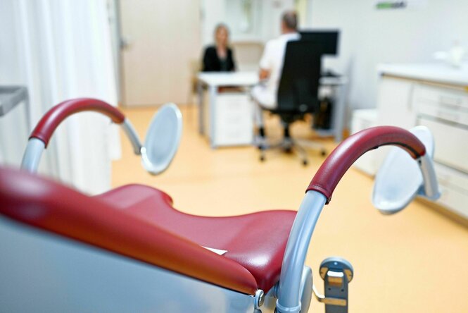 Gynäkologischer Stuhl im Vordergrund, im Hintergrund eine Konsultation zwischen medizinischem Personal und zu behandelender Person
