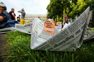 Ein Papierschiffchen aus einer Zeitung liegt auf einer Wiese, darum herum sind Menschen zu sehen
