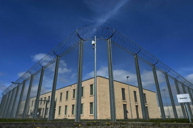 Forschperspektive, ein hoher Zaun, mit Stacheldraht bewehrt, dahinter ein weißes Gebäude, offensichtlich ein Gefängnis