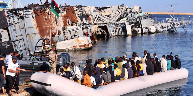 Schlauchboot mit lauter Menschen legt an, im Hintergrund ein auf der Seite im Wasser liegendes Militärschiff und die libysche Fahne