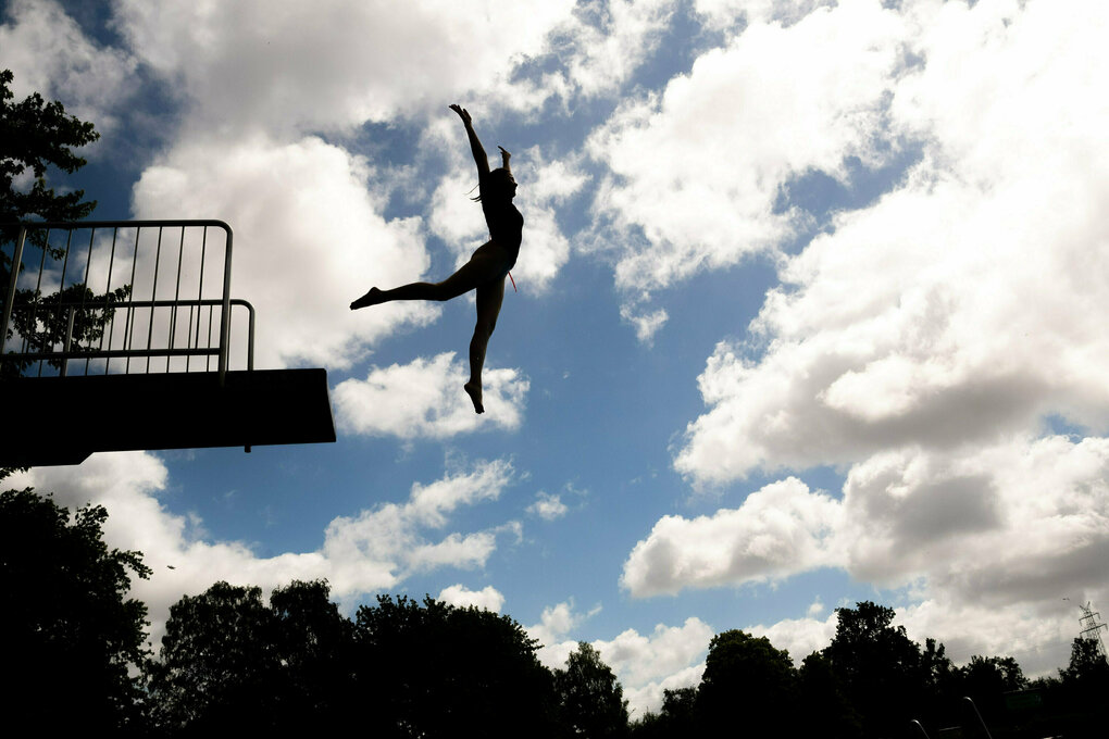 Eine weiblich gelesene Person springt von einem Sprungturm, wir sehen die Person als Silhouette, freischweben scheinbar jubelned in der Luft schweben