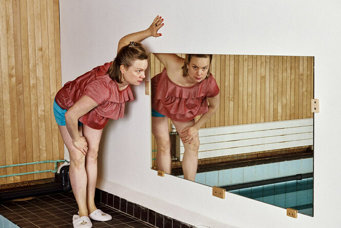 Eine Frau steht in einem holzvertäfelten Schwimmbereich und schaut in einen Spiegel. Im Spiegelbild ist ein Pool zu erkennen. Die Performerin trägt Shorts, ein glitzerndes Oberteil und Hotelschlappen.
