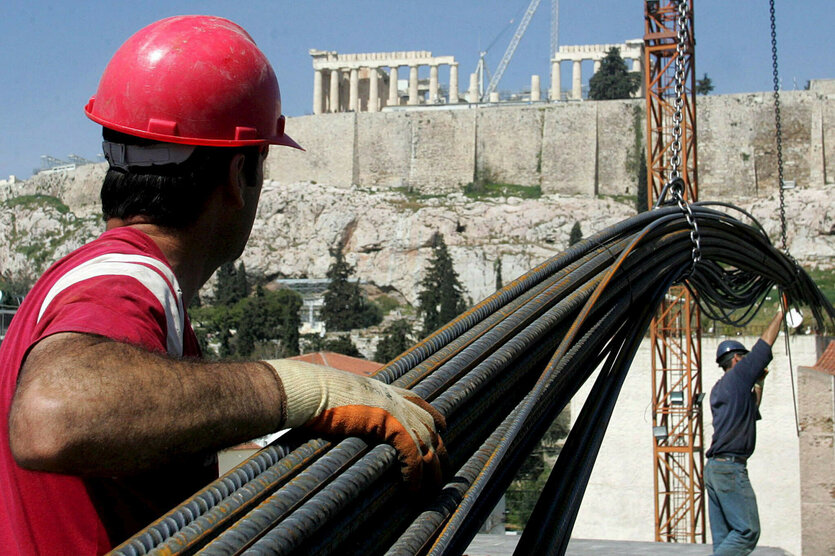 Ein Bauarbeiter trägt Stahlstangen, im Hintergrund die Athener Akropolis